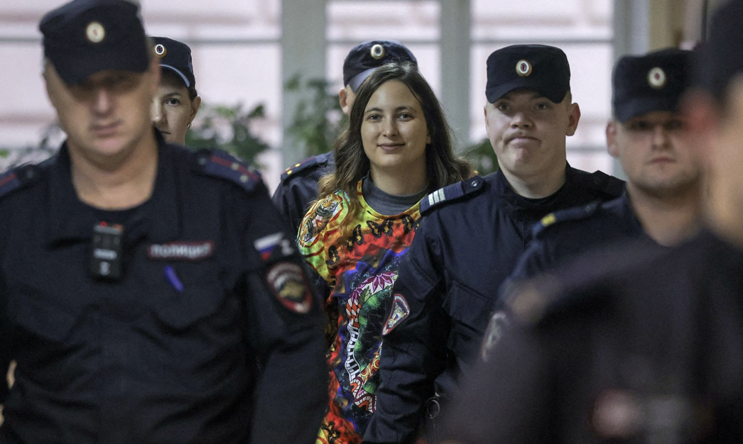 A young woman wearing a colorful shirt walks surrounded by uniformed police officers, some with serious expressions, in what appears to be an indoor setting.
